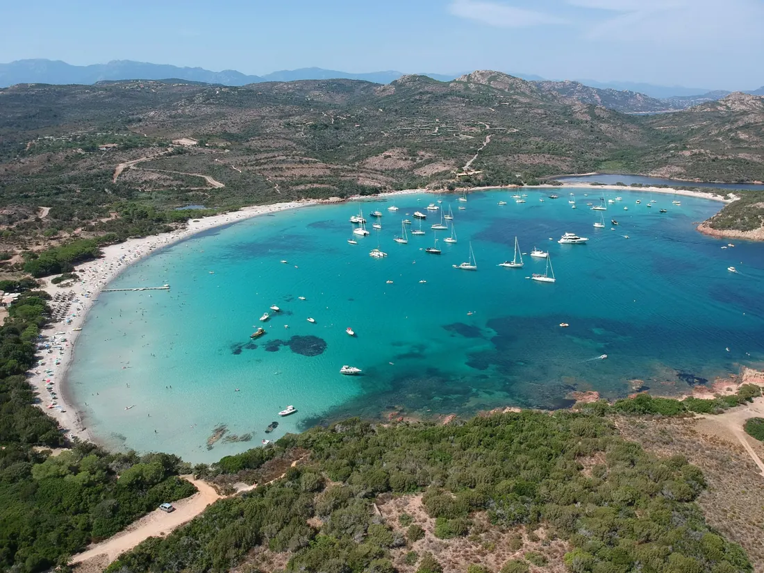 Baie turquoise entourée de maquis, avec des bateaux au mouillage